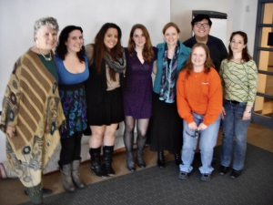 a group of eight people smiling and standing in front of a white wall. Minnie Bruce Pratt is on the far left, and Eileen Schell is the fifth person from the left.