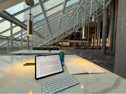 Figure 2.1: View of marble table underneath steel reinforced window in a large hotel lobby. A tablet and keyboard sit next to an open notebook on the table.